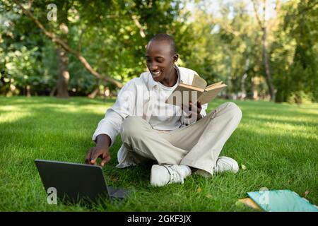 Preparing for exams outdoors. Happy black student guy studying with book and laptop computer, sitting in campus Stock Photo