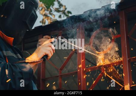 Close up of a welder hands welding steel structure metal part in home backyard. Craft man use electric arc argon wear work uniform,protective helmet o Stock Photo