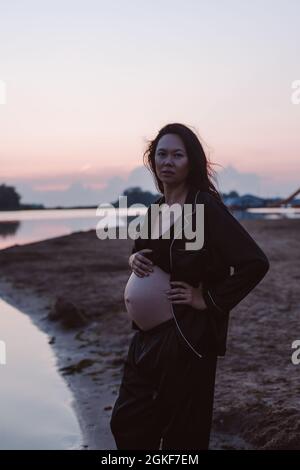 Portrait of pregnant woman on beach. Romantic photo of attractive young brunette in unbuttoned dark pajamas on beach looking at camera and her hair Stock Photo