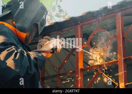 Close up of a welder hands welding steel structure metal part in home backyard. Craft man use electric arc argon wear work uniform,protective helmet o Stock Photo