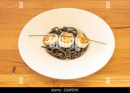 tasty black spaghetti with grilled scallops on top and an hour of chives all in a nice white plate on a wooden table Stock Photo