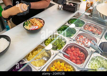Chef's gloved hands putting the finishing touches on a Hawaiian poke bowl. Metal trays with fresh ingredients Stock Photo