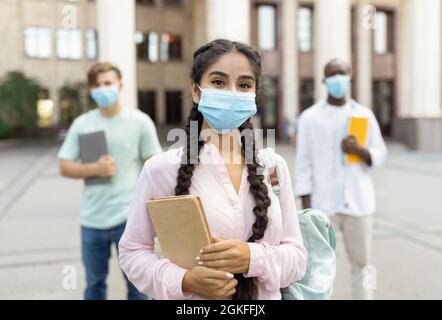 Safe study during covid-19 pandemic. Indian student girl in face mask standing with friends outdoors near university Stock Photo