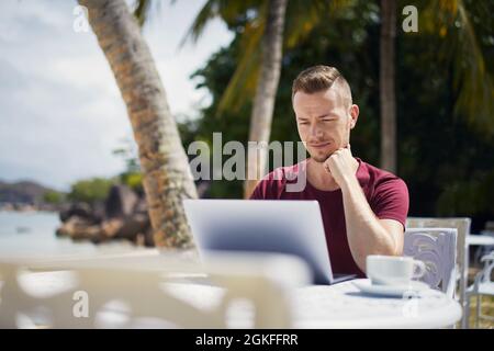 Young freelancer working on laptop from tropical destination. Man sitting under palm trees on beach. Stock Photo