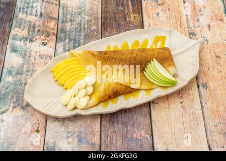 Image of sweet crepe with apple, white chocolate, mango and syrup on a white leaf-shaped plate Stock Photo