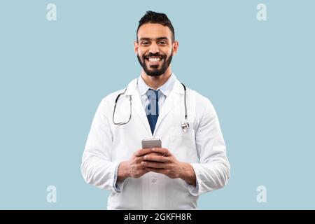 Arab Doctor Man Holding Smartphone Using Medical Application, Blue Background Stock Photo