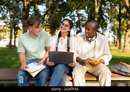 Diverse students friends learning sitting on bench, using laptop outdoors in university campus Stock Photo