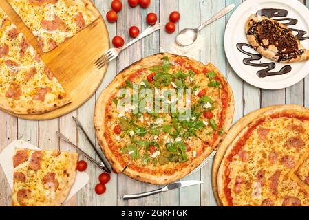 Pizza with lots of arugula, ham and york and melted cheese, sweet calzone with chocolate syrup, cherry tomatoes, knives and forks, pieces of guanciale Stock Photo