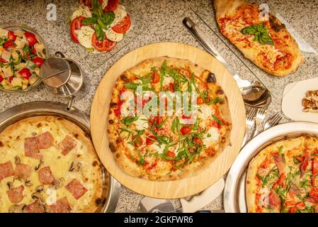 Pizzas and typical Italian dishes on a granite table. Margherita pizza with arugula, calzone with cheese, carbonara pizza with bacon, caprese salad Stock Photo