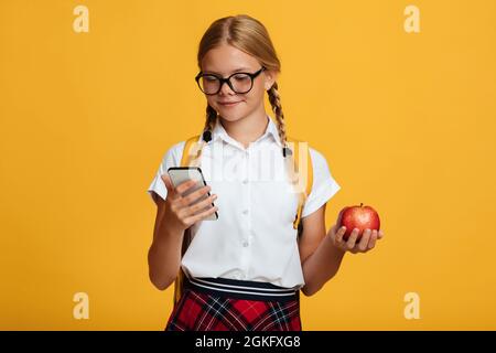 Happy young blonde girl pupil with pigtails and backpack in glasses typing on phone, holds red apple Stock Photo