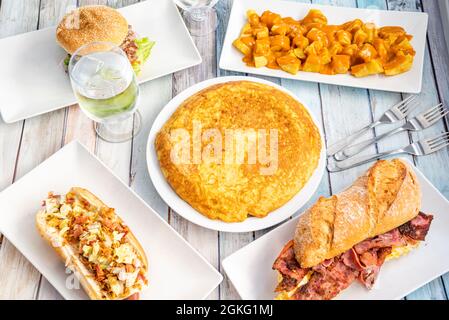 Set of Spanish food dishes with potato omelette in the center, patatas bravas, tenderloin sandwich with cheese and hot dog Stock Photo