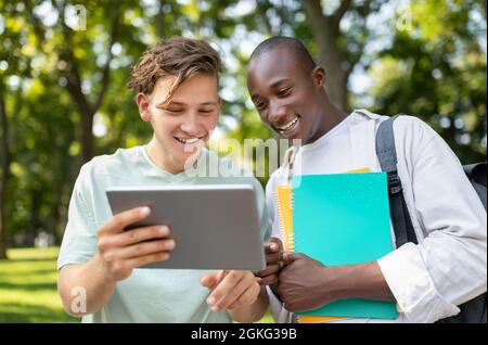 Student guy showing his project on digital tablet to friend, walking outdoors in college campus Stock Photo