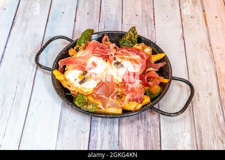 Delicious typical Spanish dish of broken eggs with serrano ham, fried peppers, homemade chips and truffle in a black enameled pan on a light table Stock Photo
