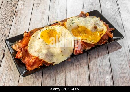 Delicious cap of broken eggs with ham and chips served in a black rectangular plate on white table Stock Photo