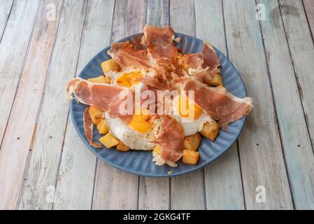 Blue plate full of fried eggs with serrano ham, diced potato chips with paprika on wooden table Stock Photo