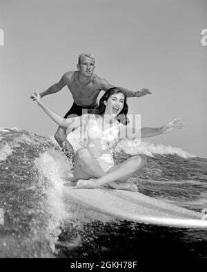 1960s ANONYMOUS COUPLE LAUGHING WOMAN SITTING ON SURFBOARD WITH MAN STANDING ARMS OUT FOR BALANCE LOOKING SERIOUS - a7692 CYP001 HARS OLD FASHION 1 FITNESS LAUGH HEALTHY YOUNG ADULT BALANCE TEAMWORK SURFING WAVES ATHLETE PLEASED JOY LIFESTYLE SPEED FEMALES MARRIED SPOUSE HUSBANDS GROWNUP ATHLETICS COPY SPACE FRIENDSHIP FULL-LENGTH LADIES PHYSICAL FITNESS PERSONS GROWN-UP MALES TEENAGE GIRL TEENAGE BOY ATHLETIC PAIRS SURF B&W MEN AND WOMEN PARTNER SKILL ACTIVITY PHYSICAL THRILL STRENGTHENING WEIGHT LOSS CHEERFUL ADVENTURE STRENGTH SELF ESTEEM EXCITEMENT LOW ANGLE RECREATION RELATIONSHIPS SMILES Stock Photo