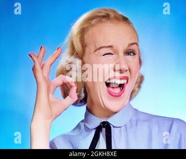 1950s ENTHUSIASTIC BLOND WOMAN MOUTH OPEN WINKING EYE LOOKING AT CAMERA AND MAKING OK SIGN WITH THUMB AND FINGER - g3462c DEB001 HARS PLEASED JOY SATISFACTION FEMALES WINNING STUDIO SHOT PORTRAITS GROWNUP HOME LIFE COMMUNICATING COPY SPACE WINNER GROWN-UP OK WIN CONFIDENCE GESTURING EXPRESSIONS EYE CONTACT BIZARRE SUCCESS WEIRD AGREE HEAD AND SHOULDERS CHEERFUL VICTORY GROTESQUE AGREEMENT EXCITEMENT LOUD OKAY LIPSTICK ZANY UNCONVENTIONAL SMILES APPROVAL JOYFUL LAUGHTER SUCCEED WACKY DEB001 IDIOSYNCRATIC OK GESTURE OK SIGN OK SIGNAL SYMBOLIC AMUSING COMMUNICATE ECCENTRIC EXCITING Stock Photo