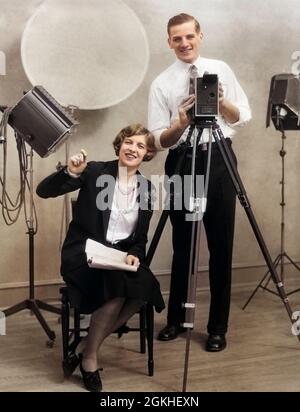 1920s PHOTOGRAPHER STANDING BEHIND MOVIE CAMERA IN STUDIO WITH ASSISTANT SITTING BESIDE HOLDING SCRIPT BOTH LOOKING AT CAMERA - o3297c HAR001 HARS NOSTALGIA MEDIA INDUSTRY OLD FASHION 1 PHOTOGRAPH PHOTOGRAPHER COMMUNICATION CAREER CREW TEAMWORK PHOTOS ASSISTANT PHOTOGRAPHS JOY LIFESTYLE LIGHTING SPEED FEMALES JOBS STUDIO SHOT DIRECTOR GROWNUP COPY SPACE FRIENDSHIP FULL-LENGTH LADIES PERSONS CAMERAS MALES CAMERAMAN PROFESSION PAIRS ENTERTAINMENT SPIRITUALITY CONFIDENCE TRIPOD MEN AND WOMEN LENS PICTURES SUCCESS TEMPTATION OCCUPATION PHOTOGRAPHERS HAPPINESS MATES MATE STRENGTH STRATEGY CAREERS Stock Photo