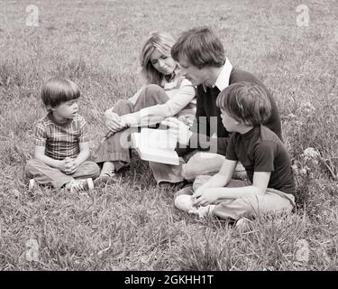 1970s FAMILY MOTHER FATHER TWO SONS SITTING IN GRASS FATHER READING TO THEM FROM A BOOK - r24365 HAR001 HARS OLD TIME NOSTALGIA BROTHER OLD FASHION 1 JUVENILE COMMUNICATION PEACE INFORMATION SONS FAMILIES LIFESTYLE PARENTING RELIGION FEMALES MARRIED BROTHERS RURAL SPOUSE HUSBANDS HOME LIFE COPY SPACE FRIENDSHIP HALF-LENGTH LADIES PERSONS INSPIRATION MALES CHRISTIAN SIBLINGS SPIRITUALITY FATHERS B&W PARTNER FREEDOM HIGH ANGLE DISCOVERY RELIGIOUS CHRISTIANITY DADS SIBLING CONNECTION HOLY FAITHFUL COOPERATION FAITH JUVENILES MID-ADULT MID-ADULT MAN MID-ADULT WOMAN MOMS SPIRITUAL TOGETHERNESS Stock Photo