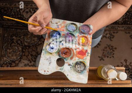 working, an artist's hand full of paint while mixing colors on a palette with a brush near oils to work, work tools, hobbie, creative lifestyle Stock Photo