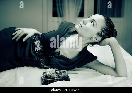 Young woman lying on a bed of white sheets looking into the light dressed in a dark vintage style dress, with an old book and a dried flower by her si Stock Photo