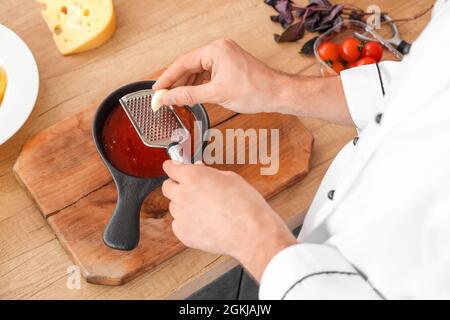 Male chef grating garlic into saucepan at table in kitchen, closeup Stock Photo