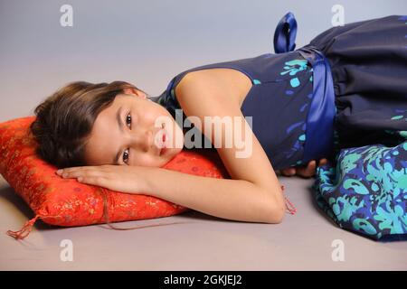 Portrait of a beautiful dark-haired teenage girl 12 years old in a blue dress with open hands. She rested her head on the red pillow and looked eye to Stock Photo