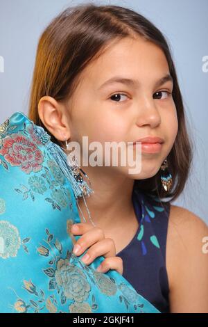 Close-up portrait of a bright cute dark-haired teenage girl 12 years old in a dark blue dress, she playfully peeks out from behind a blue silk pillow. Stock Photo