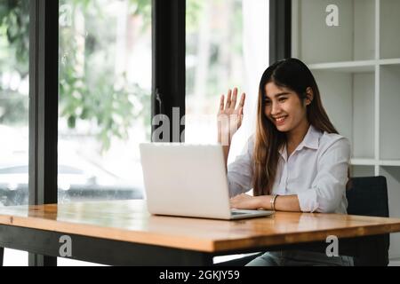 Smiling asian girl talking and waving hand hello during online video call via laptop computer, sitting in coffee shop. Cheerful business woman having Stock Photo