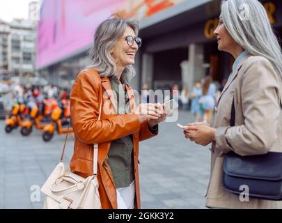 Smiling mature women and Asian lady with modern mobile phones talk on city street Stock Photo