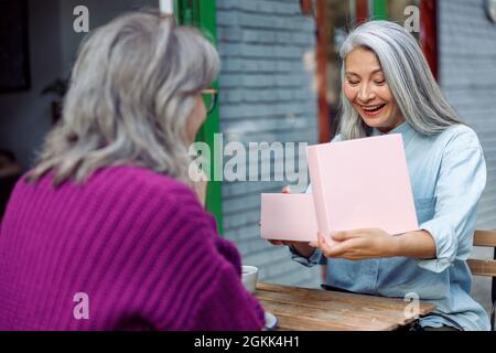 Happy senior Asian lady opens gift with best friend celebrating holiday in cafe Stock Photo
