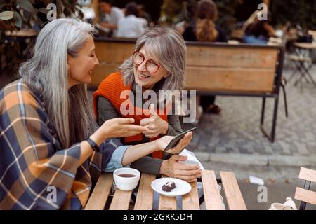 Smiling senior woman shows thumb up to friend holding cellphone in street cafe Stock Photo