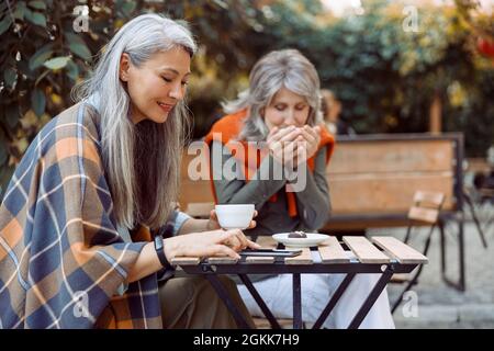 Senior Asian woman uses mobile phone while hoary haired friend drinks tea in street cafe Stock Photo