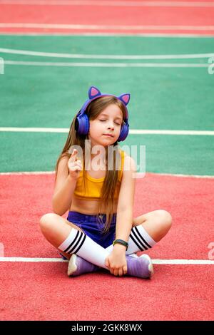 Relaxed carefree preteen girl eyes closed with wireless headphones wears trendy clothes, chilling on sports ground, enjoying song single pop rock having fun alone. Inner world. Meditation Stock Photo