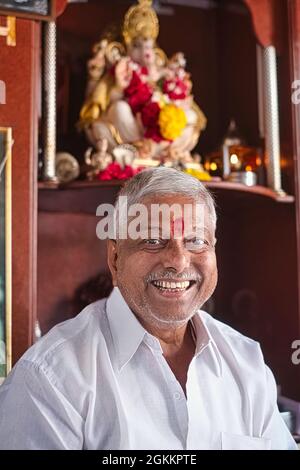 A smiling Hindu shopkeeper in Mumbai, India, with a red tika mark on his forehead and a Ganesh (Hindu elephant god) figure behind him Stock Photo