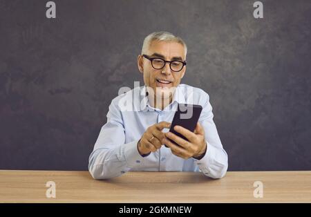 Smiling senior man writes message or browses news on Internet using modern mobile phone. Stock Photo