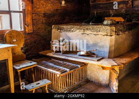 Dark wooden clogs and an iron implement on a wooden stand rest on a masonry stove bench in Museum Walserhaus (Bosco Gurin, in the 1386 building since 1938). A simple wooden slatted rack stands in front. Circolo della Rovana, Ticino, Switzerland. Stock Photo
