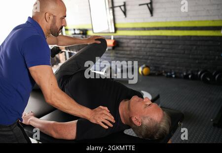 Male personal trainer and physiotherapist assisting mature man in stretching hip and leg. Assisted strength and injury recovery training Stock Photo