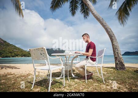 Young freelancer working on laptop from tropical destination. Man sitting under palm trees on beach. Stock Photo