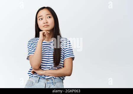 Image of asian girl thinks, looks aside with thougtful face expression, deciding smth, stands against white background Stock Photo