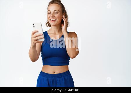 Smiling fitness woman listening music in wireless headphones. Sportswoman listening to sports instructor on smartphone app, workout in earphones Stock Photo