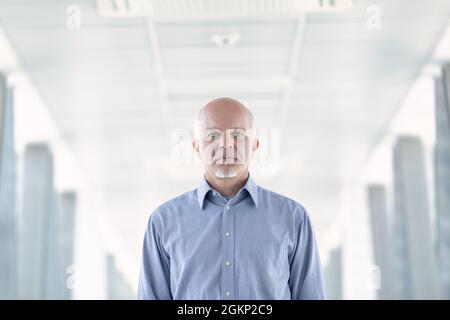 Senior businessman with a deadpan serious expression approaching the camera through a high key passageway in a commercial building in a low angle view Stock Photo