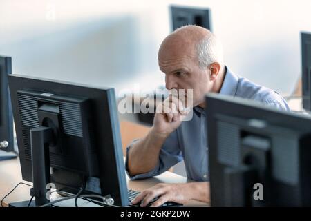 Hardworking businessman concentrating on his desktop computer leaning forwards as he reads data on the screen viewed between monitors Stock Photo
