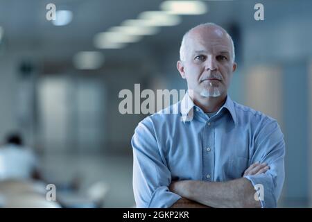 Successful confident senior business man staring thoughtfully at the camera with folded arms and a grim expression indoors in an open plan office Stock Photo