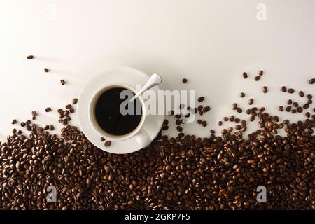 White cup of coffee on saucer on white table and pile of coffee beans on the bottom. Top view. Horizontal composition. Stock Photo