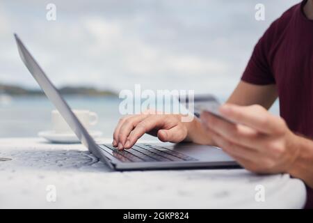 Man sitting on beach against sea and using credit card for online reservation or shopping via laptop. Stock Photo