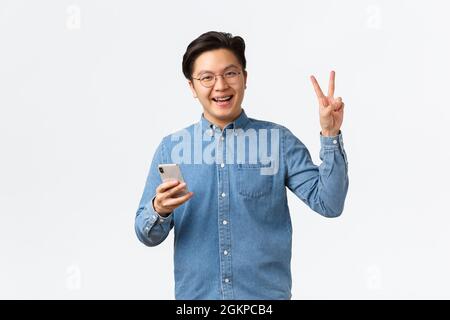 Cheerful asian man with braces, wearing glasses and shirt, using mobile phone, smartphone application, make peace kawaii gesture and smiling at camera Stock Photo