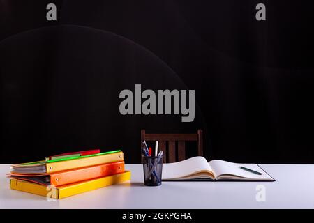 Pencils pot next to the stacked books and an open notebook , school supplies on white desk with blackboard texture in background. Side view, copy spac Stock Photo
