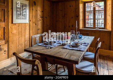 A rustic wooden table is set for dinner in the wood-panelled parlour of Cà Vegia in Cerentino, wine glasses and a candle stand ready. The patrician house, built around 1600, offers four bedrooms and deliberately does without electricity. Circolo della Rovana, Ticino, Switzerland. Stock Photo