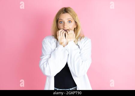 Young blonde doctor woman wearing stethoscope standing over isolated pink background looking stressed and nervous with hands on mouth biting nails. an Stock Photo